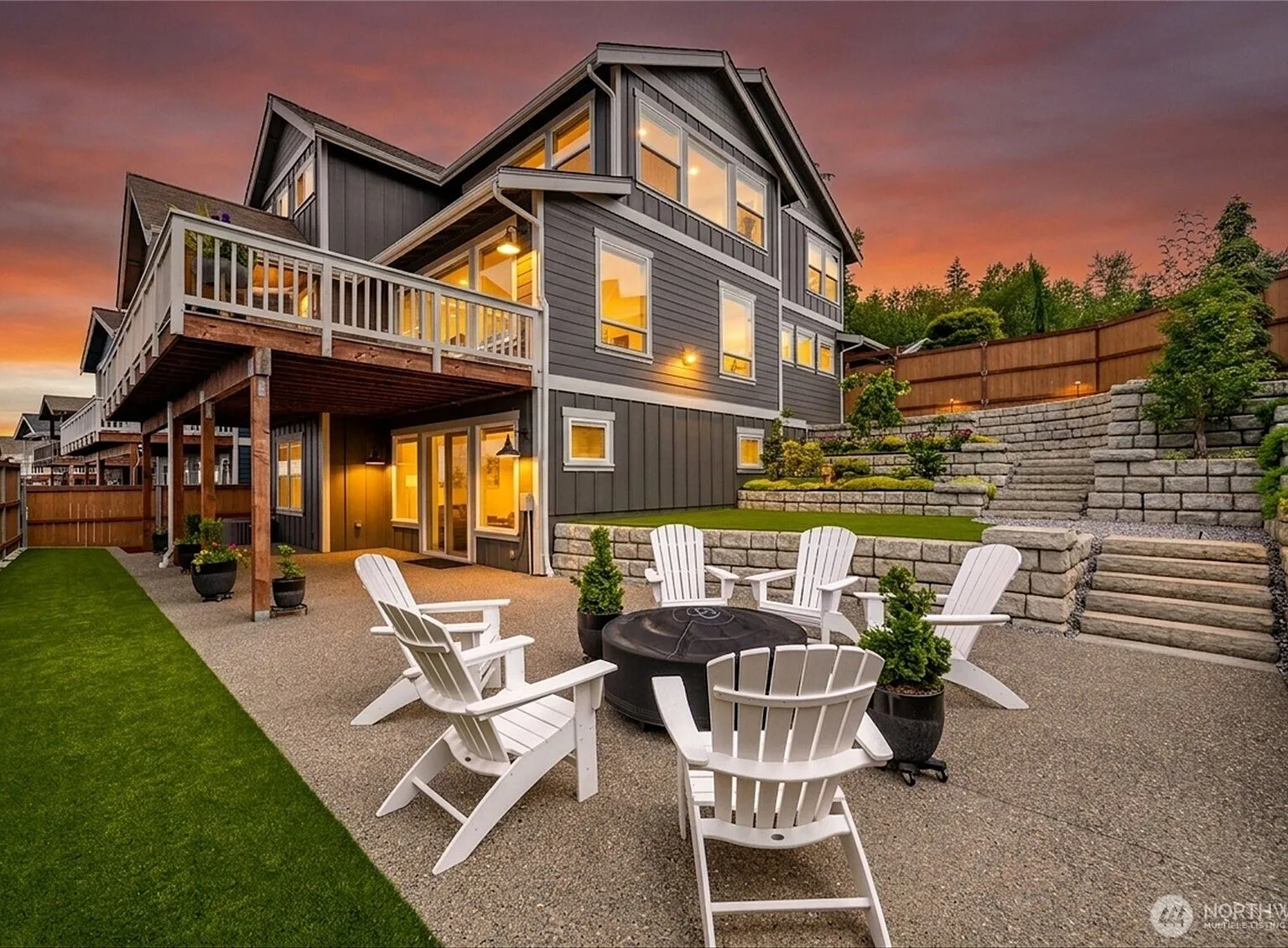 Rear patio at dusk with a fire pit and white Adirondack chairs, backed by tiered stone retaining walls and a lawn.