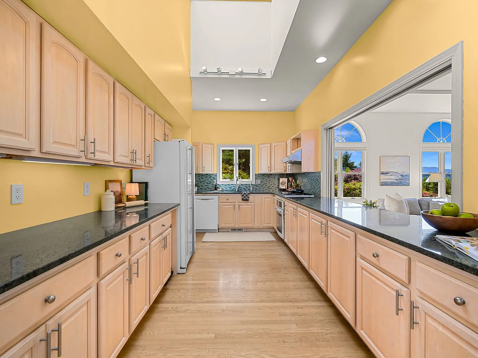 Galley kitchen with light wood cabinets and dark granite counters, opening through arched windows to the living room.